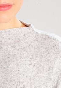Hellgrauer Pullover mit hohem Kragen, gefertigt aus weichem, strukturiertem Material mit einem subtilen Melange-Muster. Nahaufnahme des Kragens und Gesichtsbereichs.