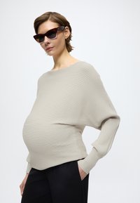 Béžový rebrovaný pletený top s voľným strihom a lodičkovým výstrihom, v kombinácii s čiernymi nohavicami. Má dlhé rukávy a textúrovaný povrch.