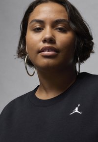 Sort sweatshirt med rundt udskæring og hvid Jordan-logo på venstre bryst. Modellen har store sølv hoopsøreringe på.