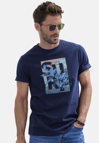 Navyblaue Baumwoll-T-Shirt mit einem Grafikdruck, der tropische Blätter, geometrische Formen und strukturierte Akzente zeigt. Umschlagärmel, lässige Passform.