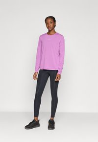 Μακρυμάνικη αθλητική μπλούζα ανοιχτό μοβ με στρογγυλή λαιμόκοψη και μικρό λευκό λογότυπο, συνδυασμένη με μαύρο εφαρμοστό κολάν και μαύρα αθλητικά παπούτσια.