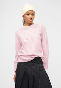 Νεαρή γυναίκα που φοράει λευκό πλεκτό σκουφάκι, ροζ και λευκό ριγέ μακρυμάνικο πουκάμισο και μαύρη τζιν φούστα με πιέτες, σε απλό φόντο.