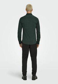 Camisa de manga larga verde oscuro con cuello estándar, yugo trasero y puños de botón, combinada con pantalones negros y zapatos negros.