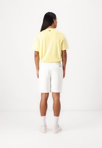 Gul kortermet skjorte med en liten logo på baksiden, kombinert med hvite shorts. Fottøyet er i lys farge, med synlige ankelsokker.