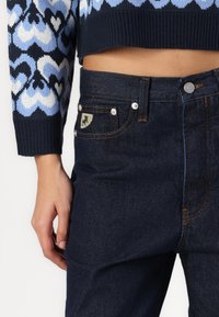 Nainen, joka on pukeutunut tummansinisiin farkkuihin ja lyhytkuituiseen neulepaitaan, jossa on sinivalkoinen sydänkuvio, käsi lepäämään hänen sivullaan.