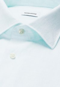 Chemise bleu clair à col pointu, deux boutons blancs et une étiquette de marque « seidensticker » visible à l'intérieur du col.