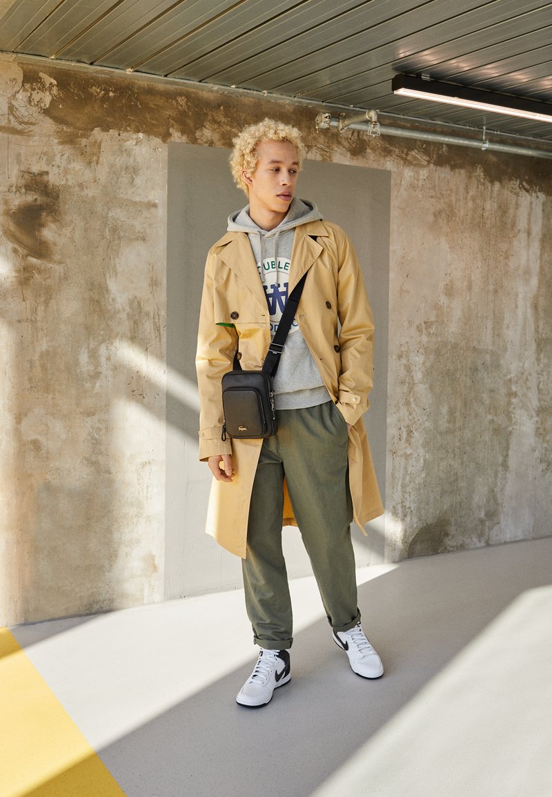 Beige trenchcoat med dobbeltknaplukning over grå hoodie, olivengrønne bukser og hvide sneakers med sorte detaljer; sort crossover taske.