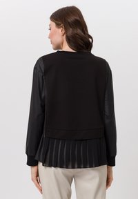 Vrouw met bruin haar draagt een zwarte long sleeve top met leren mouwen en een geplooide chiffon achterpan, gecombineerd met lichtbeige broek.
