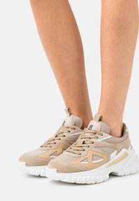 Sneakers beige con un mix di materiali in suede e mesh. Suola bianca chunky, colletto imbottito e sistema di allacciatura prominente. Design a ritaglio notevole.