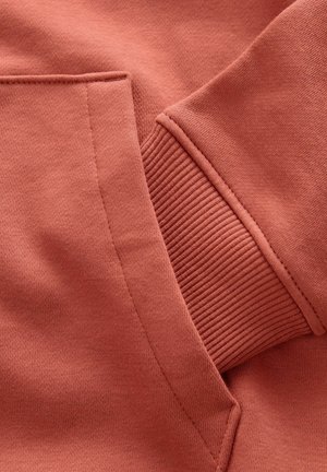 Materialul puloverului de culoare ruginie arată o textură netedă, cu un manșon ribbed și buzunar. Detaliile cusăturilor sunt vizibile, punând în evidență designul articolelor de îmbrăcăminte.