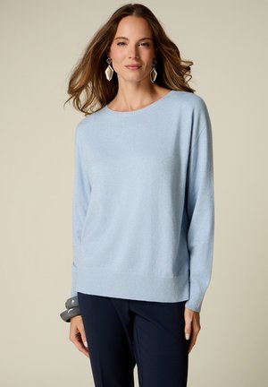 Vrouw met lang bruin haar die grote geometrische oorbellen draagt, een lichtblauwe trui, donkere broek en grijze armbanden aan haar linkerpols.