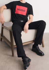 T-shirt nera con logo rettangolare rosso "HUGO", abbinata a pantaloni neri e scarpe stringate nere. Seduto su una sedia imbottita chiara.