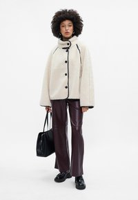 Fluffy crème jas met hoge kraag, zwarte knopen en piping; bordeaux kunstleren broek; zwarte handtas; chunky zwarte schoenen.