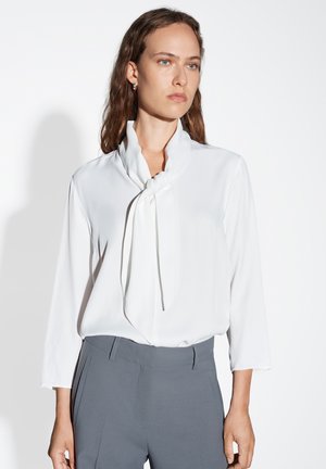 White blouse with a tie neck detail, featuring a smooth texture and long sleeves, paired with gray high-waisted trousers.