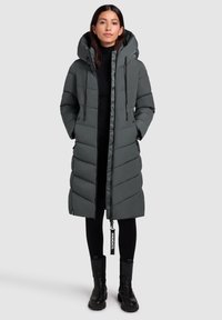 Femme portant un long manteau gris rembourré avec une capuche, des leggings noirs et des bottes noires, se tenant devant un fond gris clair uni.