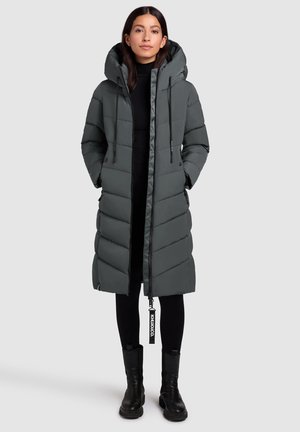 Femme portant un long manteau gris rembourré avec une capuche, des leggings noirs et des bottes noires, se tenant devant un fond gris clair uni.