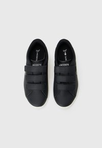 Černé kožené boty s hladkým povrchem, vybavené třemi suchými zipy, bílou podrážkou a logem Lacoste na boku a vnitřní sole.