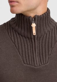 Brauner Strickpullover mit hochkanntem Rippenkragen, halbem Reißverschluss und einem Lederzugband. Verfügt über eine glatte Textur und ausgeprägte Nähte.