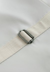 Beige vävd tygrem med ett metallrektangulärt spänne på en slät, ljus bakgrund.