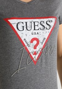 Grå v-ringad t-shirt med en röd och vit triangelgrafik som har texten "GUESS U.S.A." och rhinestones som stavar "Icon."