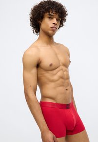 Boxers de algodão vermelhos com uma faixa na cintura com logótipo. Textura suave, design ajustado e comprimento até a meia coxa. Modelo em pose de lado.