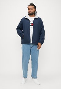 Jachetă ușoară albastru închis cu guler gri, purtată peste un tricou alb cu logo, asortată cu blugi din denim deschis la culoare și adidași albi.