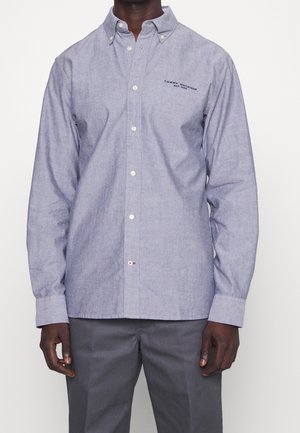 Homme portant une chemise bleu clair à boutons avec le logo "Tommy Hilfiger" et un pantalon gris foncé, debout devant un fond blanc uni.