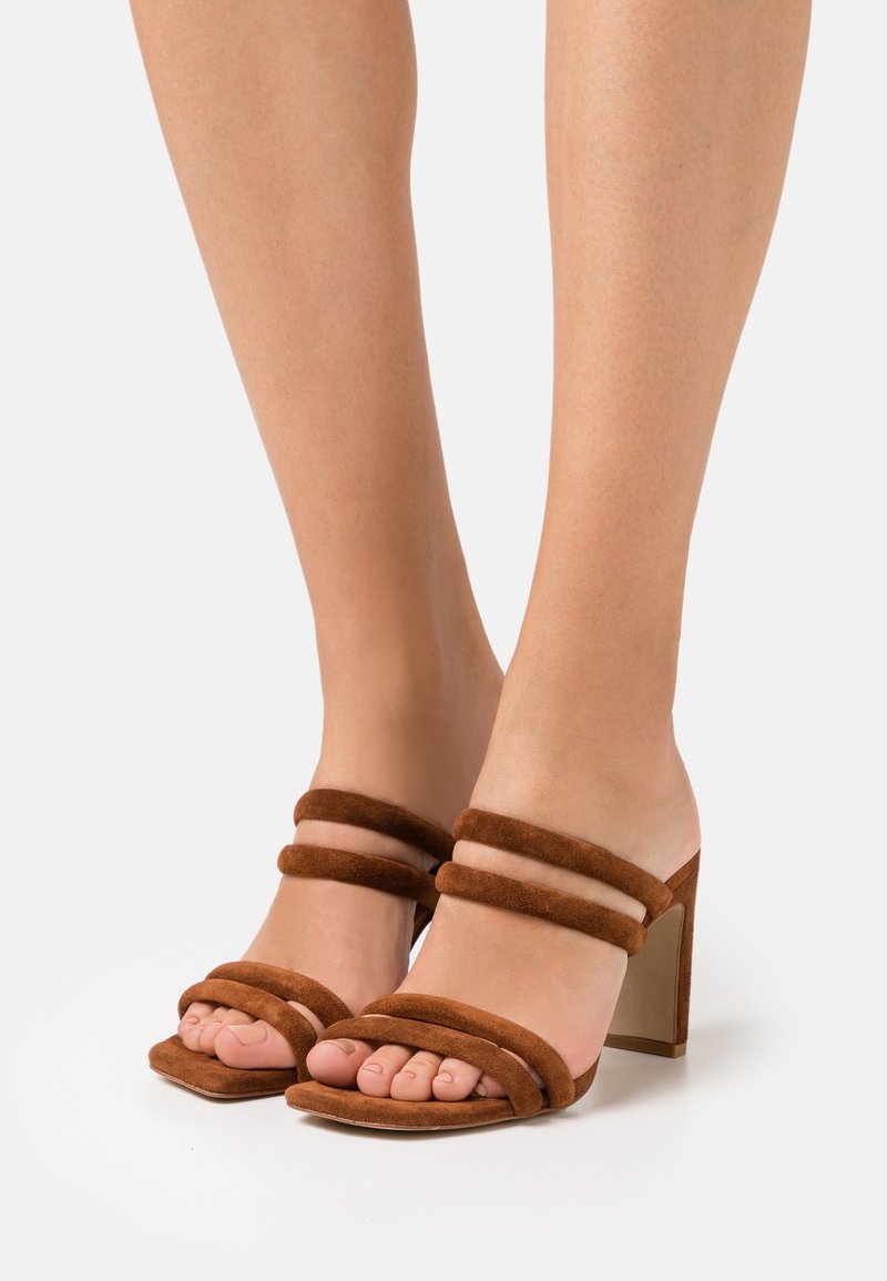 Bruine suede sandalen met hak, voorzien van drie brede banden over de tenen en een chunky hak. De zool is plat en het ontwerp is open aan de achterkant.