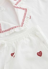 Fehér textilből készült ruhadarab elasztikus derékpánttal, piros hímzett szívekkel, fodros szegélyekkel, piros keresztszemes díszítéssel.