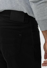 Schwarze Jeans aus strukturiertem Stoff, mit einem Lederpatch am Bund und einem kleinen geprägten Logo. Dezente Kontrastnähte.