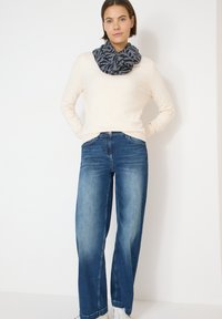 Pull à manches longues beige, jean bleu à jambes larges, écharpe noire et grise à motif, baskets blanches, design minimaliste, texture lisse.