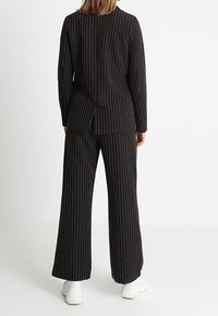 Costume à fines rayures noires avec une veste ajustée et un pantalon à jambes larges, doté d'une fente à l'arrière et de baskets blanches unies. Texture de tissu lisse.