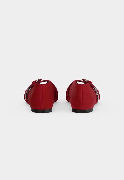 Paire de chaussures plates rouges avec des nœuds décoratifs et des accents argentés, montrée de dos sur un fond blanc uni.