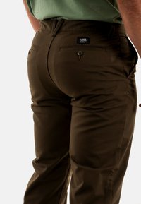 Pantaloni marroni realizzati in tessuto misto cotone con una vestibilità su misura, tasche posteriori visibili con chiusura a bottone e un'etichetta con logo sulla vita.