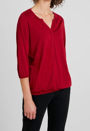 Frau mit einer roten, langärmligen Bluse mit V-Ausschnitt und schwarzer Hose, stehend vor einem schlichten hellgrauen Hintergrund.