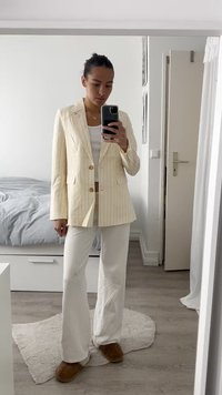 Bež blazer s prugami in zlatimi gumbi, v kombinaciji z belim vrhom in širokimi hlačami. Mehka, teksturirana tkanina in sproščen kroj.