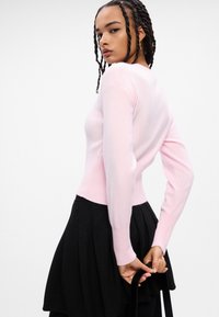 Ανοιχτό ροζ εφαρμοστό πουλόβερ με μακριά μανίκια και ριγέ στρίφωμα, συνδυασμένο με μια μαύρη πτυχωτή φούστα. Λείος υφασμάτινος σχεδιασμός, μινιμαλιστική αισθητική.