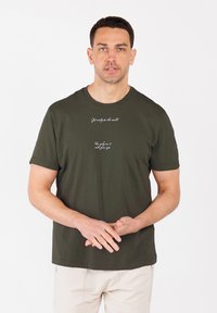 Camiseta de algodón verde oscuro con mangas cortas. Presenta texto impreso en blanco en dos líneas en el frente. Diseño simple, corte relajado, cuello redondo.