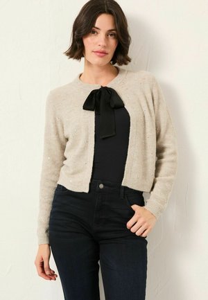 Beige gestrickte Strickjacke mit subtiler Glitzereinlage, runder Halsauschnitt und schwarzem Schleifendetail am Kragen, kombiniert mit einem eng anliegenden schwarzen Oberteil und dunklen Jeans.