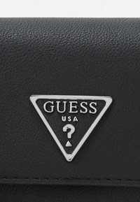 Svart plånbok i syntetläder med en silvertriangulär logotypplatta med texten "GUESS" och en frågeteckendesign. Texturerad yta.