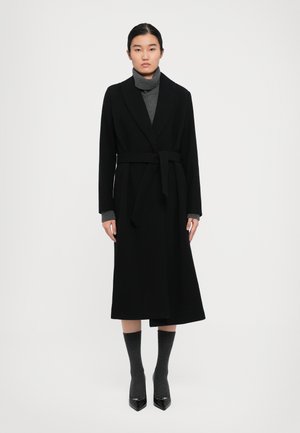 Frau in einem langen schwarzen Gürtelmantel über einem grauen Rollkragenpullover, grauen Socken und schwarzen spitzen Pumps, die vor einem weißen Hintergrund steht.