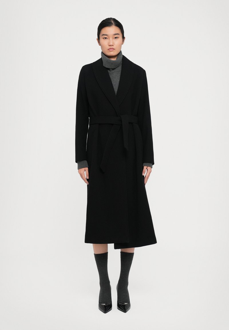 Woman wearing a long black belted coat over a gray turtleneck sweater, gray socks, and black pointed-toe heels, standing against a white background.