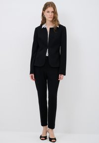 Black tailored blazer with a round neckline and two front pockets, layered over a white shirt with a black polka dot pattern and black fitted trousers.