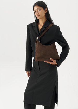 Woman wearing a black blazer and skirt, holding a large dark brown suede shoulder bag against a white background.