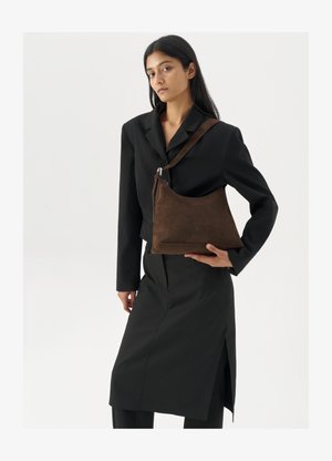 Kvinna som bär en svart kavaj och kjol, hållande en stor mörkbrun mocka axelväska mot en vit bakgrund.