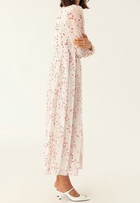 Vestido largo y fluido hecho de una tela blanca transparente con delicados patrones de hojas rojas. Presenta un escote con volantes y mangas largas semi-transparentes.