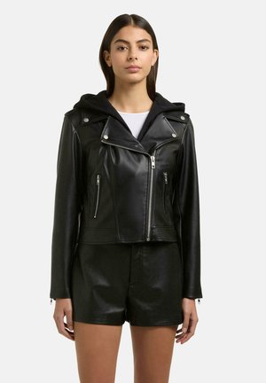 Jeune femme portant une veste de motard en cuir noir avec une capuche et un short en cuir noir assorti sur fond blanc.