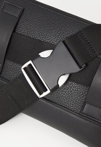 Černá taška s texturovaným koženým tělem, odolným nylonovým popruhem a kovovým uzávěrem, zdůrazňující funkční designové prvky.