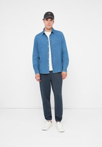 Tânăr purtând o șapcă neagră, cămașă albastră cu nasturi peste un tricou alb, pantaloni de jogging închisi la culoare și adidași albi, stând în fața unui fundal alb simplu.