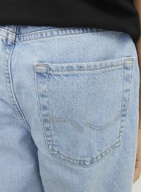 Šviesiai mėlynos džinsinės kelnės su užpakaline kišene, kuri turi subtilų kreivą siūlę. Medžiaga turi būdingą džinsinės tekstūros ir apdailos charakteristiką.